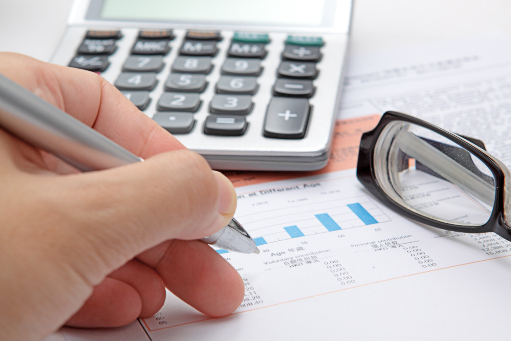 person writing on documents with charts and graphs with calculator and eyeglasses on the side